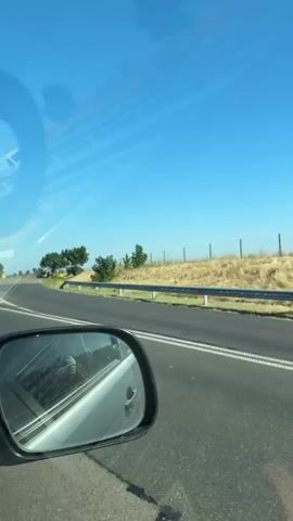The video is filmed from the perspective of a moving vehicle, likely a car, looking out the passenger side window. The road is paved with white lane markings visible. On the left side of the road, there is a shoulder and then a grassy embankment with some trees. A group of people, some wearing high-visibility vests, are standing on the shoulder or in the grassy area. Further into the background, there is a fence running along the top of the embankment, and beyond that, a field of dry, yellow grass or crops under a clear blue sky. The video continues to show the passing landscape, with more fields and trees visible. A silver car is seen in the distance on the right side of the road. The video captures a roadside scene with what appears to be some form of official presence or activity.
