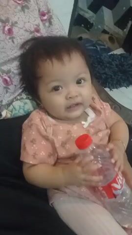 A baby is seated on someone's lap, holding a plastic water bottle. The baby is wearing a pink shirt with a floral pattern and a white ribbon. The baby has dark hair and is looking towards the camera. The background is blurred but appears to be an indoor setting with furniture. The lighting is dim.