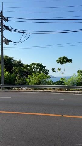 The camera moves along a highway, past an open view. Trees and bushes are on the other side of a metal guard rail, leading down to a blue ocean view. Mountains and buildings are in the background. There is a light pole and electrical wires in the foreground. The road has a yellow line on the edge, and white lines. The sky is clear and blue.