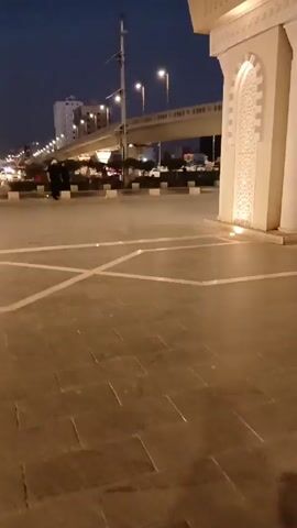 A low-angle, eye-level shot depicts a woman in a black robe walking across a paved square. The square has light-colored lines and designs. In the background, there's a city scene with buildings, streetlights, and an elevated highway. The sky is a deep blue, suggesting it's either dusk or dawn. To the right of the frame, there's a building with ornate decorations on its white façade. The camera remains stationary throughout the short clip.