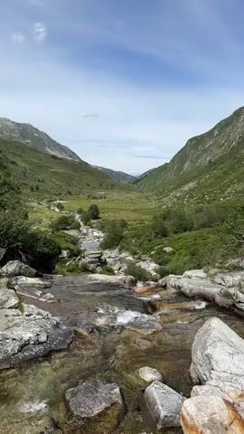The video shows a slow-moving river flowing through a mountainous valley. The foreground features rocks and shallow water with visible ripples. The midground consists of the river meandering through a green valley dotted with bushes and small trees. In the background, the valley narrows and is flanked by steep, verdant mountainsides under a partly cloudy blue sky. There are no people or animals visible in the video.