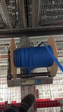 The video shows a person's foot wearing a black sock with a grey stripe near the top, standing on a silver metal structure. The person's foot is positioned in the lower left corner of the frame. In the center of the frame, a large wooden spool of bright blue cable is mounted on a silver metal stand. The cable is being unwound from the spool and routed upwards and to the right, disappearing into a network of metal racks and cable management systems. The environment appears to be a data center or a similar industrial setting, characterized by extensive metal shelving, conduits, and a grid-like ceiling structure. The background is filled with similar racks and cabling, with some areas blurred due to motion. The lighting is bright and functional, typical of such facilities.