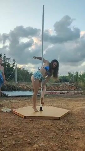 Two women are in an outdoor setting with a pole dancing apparatus. The woman on the right is performing a pole dance move, while the woman on the left observes. The ground is covered in dirt and gravel, and there is a fence in the background. The sky is cloudy.