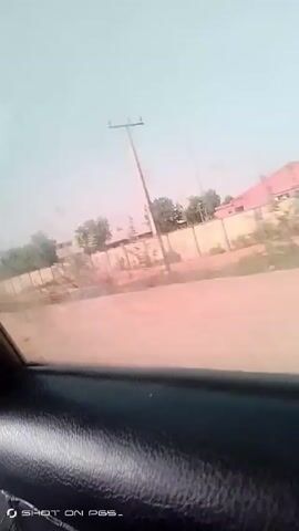 The video shows a view from inside a moving vehicle. Outside the window, a beige wall runs along a road. There are some trees and buildings visible behind the wall. The sky is pale. A utility pole stands along the roadside. The video is slightly shaky due to the movement of the vehicle.  A logo that says "SHOT ON PES-" is at the bottom of the screen.