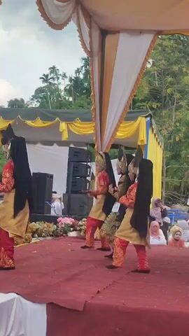 The video captures a group of individuals performing a traditional dance on a raised red stage. The performers are dressed in ornate, brightly colored costumes featuring red pants with gold trim, gold tunics, and black veils or hair extensions. They are also wearing elaborate headpieces. The stage is covered with a red carpet and decorated with white fabric along its front edge. A large tent with yellow and white fabric drapes provides shade over the stage. In the background, there are speakers, floral arrangements, and a crowd of spectators, some of whom are also wearing headscarves. Beyond the tent, lush green trees and foliage are visible, suggesting an outdoor setting. The dancers are in motion, with their poses and movements indicating a choreographed performance.
