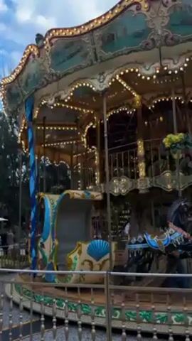 The video shows a two-story carousel spinning slowly, lit by strings of small, warm lights. The carousel is painted in shades of cream, teal, and gold, with detailed carvings and paintings around its edges. There are a few riders visible on the top level, including children. The horses on the carousel are painted in various colors and appear to be stationary. The carousel is surrounded by a metal fence and is set in an outdoor area with trees visible in the background. The sky is overcast, suggesting it is daytime or early evening. In the distance, a few people can be seen walking, adding to the ambiance of a public park or amusement area.