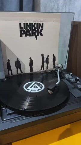 The video shows a vinyl record player with a record from the band Linkin Park titled "Minutes to Midnight" on it. The album cover is visible behind the record. The record is black with a white label in the center. The turntable is black and has a tonearm with a stylus resting on the record. The background is a textured gray wall. The overall scene is well-lit and the focus is on the record player and the album.