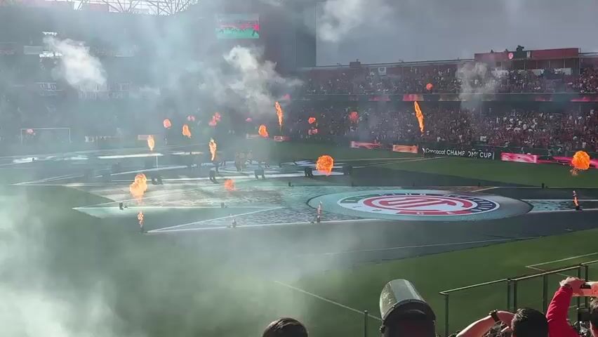 The video shows a crowded soccer stadium filled with fans and a cloud of smoke. On the field, a large, circular logo is visible. Several flames are also seen on the field, burning amidst the smoke. People in uniform are standing on the field. The atmosphere suggests a pre-game ceremony or celebration.