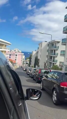 A first-person perspective shot from inside a vehicle shows a line of cars parked along a narrow road. Buildings line both sides of the street, and in the distance, the ocean is visible under a blue sky with scattered clouds. The camera focuses on the cars in front, and the side mirror shows the driver's reflection. The wind is audible.