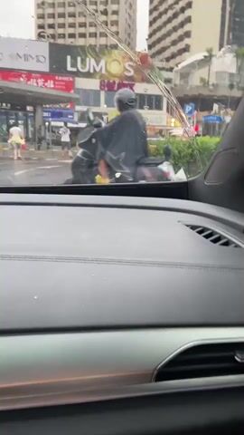 The video shows a view from inside a car, looking out through the windshield. It appears to be raining, as there are streaks of water on the windshield. Outside, a street scene unfolds. A person on a motorcycle, wearing a black cape and a helmet, is visible in the left foreground, moving away from the camera and turning right. To the right of the motorcycle rider, another person on a motorcycle, also wearing a helmet and a black jacket, is seen from the side. In the background, there are buildings, including one with a large illuminated sign that says "LUMO". There are also what appear to be bus stops or shelters with people standing underneath them. A person in a white shirt and yellow pants is walking in the middle ground. The overall impression is an urban environment experiencing rainy weather.