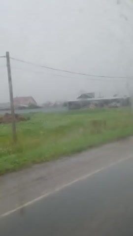A person is driving a vehicle on a wet road during a rainstorm. The camera is positioned inside the vehicle, looking out the windshield. Raindrops are visible on the windshield, distorting the view of the outside scenery. The landscape consists of green grass, a few houses, utility poles, and a dirt road. The sky is overcast and hazy, typical of rainy weather. The video captures the movement of the vehicle as it travels along the road.