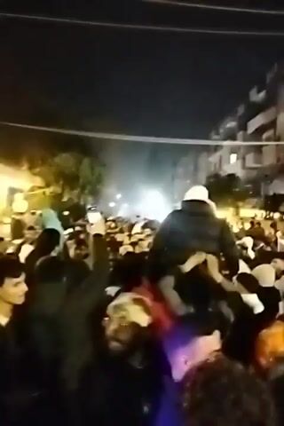 At night, a dense crowd of people is seen celebrating. The focus is on the central figure who is carried on someone's shoulders, wearing a white hat and a dark jacket. Many in the crowd are holding up their phones, likely to capture the moment. The lighting is uneven, with some areas well-lit and others darker, creating a sense of depth. Buildings line the background. The general atmosphere appears festive and lively.