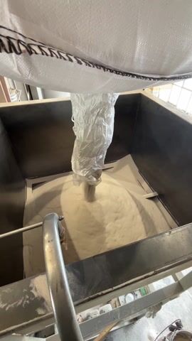 The video depicts an industrial process of bulk material handling. A large, white woven polypropylene bulk bag (FIBC) is suspended by its black reinforced lifting straps above a substantial, rectangular metal container or hopper.

A light-colored, finely powdered or granular material flows steadily from the discharge spout at the bottom of the bulk bag, through a clear plastic liner, and into the metal container below. The material accumulates in the container, forming a conical pile. A curved metal pipe or fixture is visible within the lower left portion of the container.

The setting is an indoor industrial facility, characterized by overhead yellow structural beams and ambient daylight entering from a window in the background, indicating it is daytime. No people are visible in the video. No discernible text appears on either the bulk bag or the container. The activity showcases a controlled transfer of dry bulk goods in a manufacturing or warehousing environment.