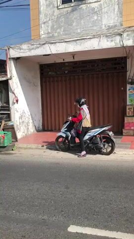 A person wearing a helmet, hijab, and a white and red jacket sits on a white and blue scooter parked on the side of a street. The scooter is facing right. The person is holding a black bag with a red strap. Behind the scooter is a building with a large, corrugated metal roll-up door that is closed. To the left of the door, there is a white wall that is peeling and weathered. A green plastic crate with some items is visible to the far left of the building. The street in front of the scooter is paved. The lighting suggests it is daytime.