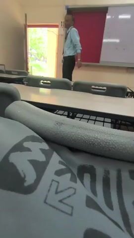 The video shows a classroom setting. In the foreground, a close-up shot of a grey fabric with a white graphic design is visible. In the background, a man in a light blue shirt and dark pants stands near a doorway, facing away from the camera. A whiteboard with some writing is on the wall behind him. Another person, also wearing a light blue shirt and dark pants, walks into the frame from the right and stands near the first man. Both men then walk towards the doorway and exit the frame. The classroom appears to be empty otherwise.