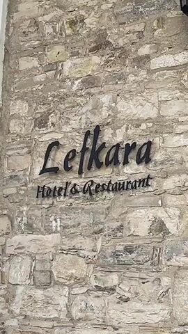 The video shows the exterior of a hotel and restaurant called Lefkara. The building has a stone facade and a wooden door with glass panels. The words "Lefkara Hotel & Restaurant" are displayed on a sign above the door. There is a table and chairs outside the entrance, as well as two black lanterns mounted on the wall. The video pans slightly to the right, showing more of the building's exterior and the entrance to the restaurant.