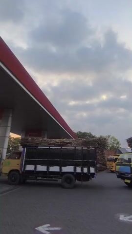 A yellow and blue truck carrying sugarcane stalks is parked at a gas station. The truck is loaded with a large amount of sugarcane. The gas station has a red and white roof. The sky is cloudy. Another truck, loaded with sugarcane, is visible in the background.