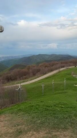 The video shows a series of large, metallic sword sculptures on a grassy hillside overlooking a mountainous landscape. A road winds through the hills in the distance. The sky is cloudy with some patches of light. The camera moves to show different angles of the swords and the surrounding scenery.