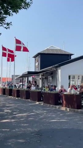 The video captures an outdoor seating area of a restaurant on a bright, sunny day. Two Danish flags fly prominently on flagpoles to the left of the frame. The restaurant has a white exterior with a dark roof structure and outdoor seating. Several people are seated at tables, some appearing to be engaged in conversation. A person walks from left to right across the foreground of the video. The ground is paved and appears to be asphalt, with dappled shadows from trees falling across it. In the background, there are boats and masts, suggesting the location is near a harbor or marina.