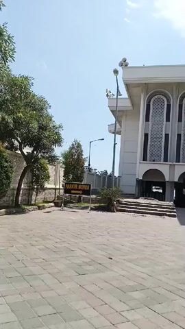 The video shows an outdoor scene in front of a building with a white facade and decorative arched windows. A paved area with square tiles is in the foreground, with a yellow trash can visible on the right side. A sign reading "PARKIR MOTOR" (Motorcycle Parking) is posted on the left. Several people are sitting and standing near the entrance of the building, which features arches supported by dark columns. A motorcycle is parked nearby, and a person is sitting on it. The sky is clear and blue, indicating a sunny day. Trees with green foliage are present, with one large tree prominently featured on the right side of the frame.