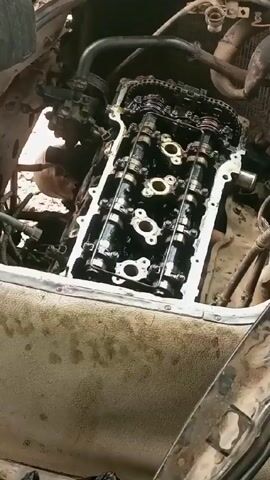 The camera focuses on an engine with its valve cover removed, revealing the camshafts and timing chain. The engine bay is dusty and appears to be in a vehicle undergoing maintenance or repair. A hand briefly enters the frame near the bottom right, then a phone is heard ringing, but not seen. The background shows additional engine components and wires.