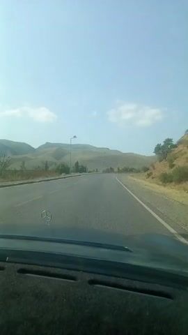 The video begins with a view from the inside of a car looking out through the windshield. The car appears to be driving on a paved road that curves to the right. The landscape outside consists of rolling hills with sparse vegetation, predominantly dry grass and some scattered trees. The sky is clear with a few scattered white clouds, indicating fair weather. Several streetlights are visible along the side of the road. The hood of the car, with a Mercedes-Benz emblem, is visible in the foreground. The overall impression is of a scenic drive through a hilly, possibly arid, region.