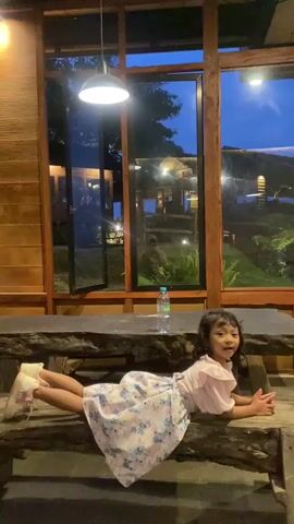 A young girl is lying on a rustic wooden table or bench inside a building, possibly a restaurant, in front of a large window that looks out onto an outdoor area with visible structures and greenery under a blue sky at dusk. The girl is wearing a light pink dress with a floral pattern and white sneakers. She is lying with her legs spread apart and her feet raised slightly, resting on the bench. Her arms are extended in front of her, supporting her upper body. She is looking directly at the camera and smiling slightly. A bottle of water is visible on the table near her. The interior of the building appears to be paneled with dark wood, and a pendant light hangs from the ceiling above the table. The outdoor area visible through the window shows other buildings with lights on, indicating it is getting dark outside.