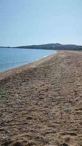 The video shows a view of a beach with the ocean and sky. The foreground is the sandy beach, covered with small pebbles and shells. The ocean is to the left, with clear blue water gently lapping at the shore. The sky is a clear, bright blue. In the distance, there is a coastline with hills. The camera is panning slowly to the right. There are no people or any other objects visible in the video. The overall impression is one of a peaceful and serene beach scene.