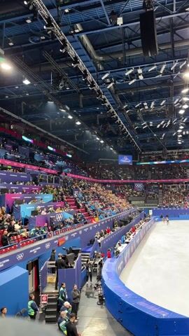 The video shows an indoor ice rink packed with spectators. A large screen suspended from the ceiling displays a video of two figure skaters, followed by a group of five individuals. Two skaters are visible on the ice. The building has a blue ceiling structure with many lights. A blue barrier surrounds the ice rink. The seating is tiered and filled with people wearing various colors. There's a small platform on the left side of the rink.