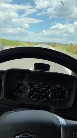 The video shows a first-person perspective of someone driving on a highway. The dashboard of the vehicle is visible, indicating a speed of approximately 90 km/h. The person driving is wearing a metal watch. The side mirror reflects the highway behind the vehicle, with other cars visible in the distance. The sky is partly cloudy.