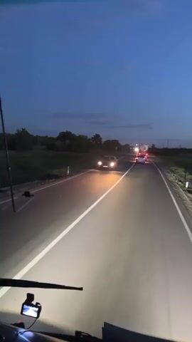 The video captures a nighttime drive on a highway in Baiqadam, Kazakhstan. The perspective is from inside a vehicle, likely a bus or truck, with the dashboard and windshield wipers visible in the foreground. The road is paved with distinct white lane markings that are illuminated by the vehicle's headlights, creating bright beams that cut through the darkness.

The main subjects on the road are vehicles. A dark sedan is seen in the middle ground, its headlights also on, suggesting it might be stopped or moving slowly. Further ahead, several other vehicles, including what appear to be trucks, are visible with their taillights glowing red. The road itself is the primary setting, a two-lane highway stretching into the distance. The surrounding environment is characterized by a dark, rural landscape with fields or grassy verges on either side of the road. Silhouettes of trees and vegetation are visible against the twilight sky.

The time of day is clearly evening or night, as indicated by the darkness and the use of headlights and taillights. The sky is a deep blue, with a few wispy clouds. The weather appears clear, with no visible rain or fog.

The activity taking place is simply driving on a highway at night. There are no people visibly interacting with each other or with the vehicles. The mood conveyed is one of transit and quiet observation of the passing scenery. The focus is on the journey and the road ahead.

There is no visible text in the media file.