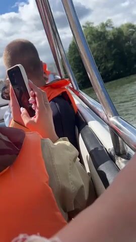 A handheld video shows a person holding a smartphone with a cracked screen while sitting on a boat. They are wearing an orange life vest and have their nails painted. Another person with a short haircut, also wearing an orange life vest, is visible in the background. The boat is moving on a body of water, creating a wake. Trees and a blue sky with clouds are visible on the shore in the distance.