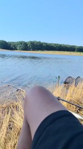 The video shows a person sitting by a lake, facing away from the camera. The person's legs and knee are visible in the foreground, with a black t-shirt or shorts. Two fishing rods are set up by the water's edge, with the lines in the lake. The lake is calm, with a clear blue sky overhead and a line of trees in the background. The ground is covered in dry grass. The scene appears peaceful and suggests an activity like fishing.