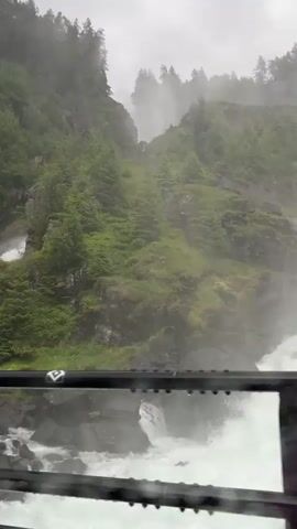 The media file captures a scenic view of a powerful waterfall cascading down a lush, green mountainside. The scene is dominated by the rugged terrain, with trees clinging to steep rocky slopes, creating a wild and untamed natural landscape. The waterfall itself is the focal point, with white water churning and crashing over rocks, generating a significant amount of mist that partially obscures the upper parts of the falls and the surrounding mountains.

In the foreground, a dark metal railing is visible, suggesting that the viewer is likely on a bridge or a viewing platform overlooking the river and waterfall. The river below the falls is also turbulent, filled with dark rocks and white, frothy water. The overall weather appears to be overcast or misty, with a soft, diffused light and a noticeable dampness in the air, evidenced by the water droplets on what appears to be a window or transparent barrier in front of the railing.

The location context of Skare, Norway, aligns with the imagery of a dramatic waterfall in a mountainous, green environment, characteristic of many Norwegian landscapes. The time of day is not explicitly clear due to the overcast conditions, but the lighting suggests it could be daytime. There are no people or discernible objects other than the natural elements and the man-made railing. The overall emotion evoked by the scene is one of awe and the raw power of nature. No visible text is present in the media file.