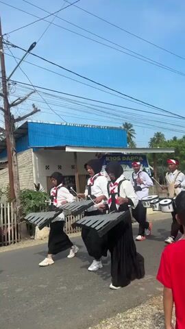 This is a vertical, shaky, outdoor video taken during the day, likely in a residential or semi-rural area with a paved road. The sky is blue with some wispy clouds. A group of young people, dressed in matching white shirts, black skirts or pants, and headscarves for some, are marching in what appears to be a parade or performance. They are carrying musical instruments. The first few individuals in the foreground are holding xylophones. Following them are several individuals playing drums of various sizes, and further back, some are playing cymbals. They are all walking in the same direction, from left to right in the frame. One individual in a camouflage uniform is walking amongst them, facing away from the camera. A few buildings, including one with a blue roof and white walls, are visible in the background, along with power lines and trees. A motorcycle is also briefly visible on the left side of the frame, with a rider. The overall impression is a community event or school performance.