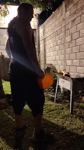 A person wearing a tank top and cropped pants approaches a grill in a yard at night. The person carries an orange plate. The grill is set up next to a brick wall and has coals burning inside. The yard is grassy and partially lit by an overhead light.