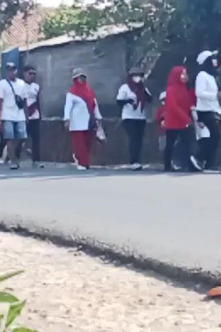 A group of people, some wearing red and white, walk along a paved road. The people appear to be participating in a parade or procession. The environment is outdoors, with a building and trees visible in the background. The footage is shaky, suggesting it was taken by someone walking or moving.