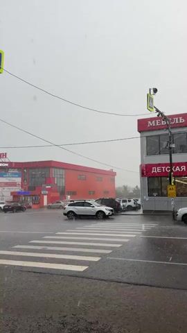 The video shows a street scene during a heavy rain shower. The road and crosswalk are wet and reflect the light. Several cars are on the road, some are moving and some are stopped. In the background, there are two buildings, one is red and the other is gray with red accents. The gray building has signage that says "МЕБЕЛЬ" and "ДЕТСКАЯ". Traffic lights are visible, and one is yellow, indicating caution. The sky is overcast, and the rain is visible as streaks falling from the sky. There are no people clearly visible in the video.