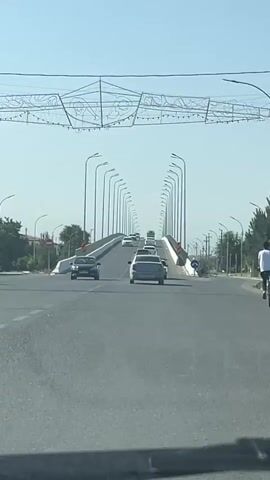 A drone view captures a wide, paved road with a bridge spanning over it. Several cars are driving on the road, with a traffic light visible. The sky is clear and blue, and trees line the sides of the road. The bridge is lined with lampposts.