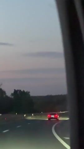 The video captures a view from inside a moving vehicle, looking forward through the windshield at a highway at dusk or early night. The sky is a muted purplish-gray, with the horizon line defined by distant dark trees or land.

On the multi-lane road, white lane markings are clearly visible. Several cars are ahead and approaching, primarily seen by their illuminated headlights and taillights. One car, in particular, is visible in the distance with red taillights. Another vehicle approaches from the opposite direction, its bright headlights creating lens flares and streaks of light on the camera at approximately the 0:02 mark, momentarily obscuring the view before passing.

The scene depicts continuous travel on the highway in Ub Municipality, Serbia, with the artificial lights of vehicles contrasting against the fading natural light of the evening. No specific people or discernible text are visible.