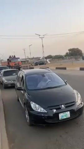 The video shows a stationary black Peugeot car in the foreground, with other vehicles behind it. The Peugeot has a license plate that reads "GWA 40DJ". The camera pans to the right, showing an orange dump truck passing by. There are several people sitting on top of the truck's cargo bed. The truck is driving away from the camera. The environment appears to be an urban or suburban road with streetlights and other vehicles. The sky is a pale blue, indicating either early morning or late afternoon.