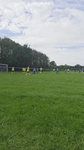 A wide shot shows a soccer game on a grassy field under a cloudy sky. Several players in yellow and blue uniforms are visible, spread across the field. Some players are running, while others are standing or kicking a ball. A blue line marks the boundary of the field. Trees line the far edge of the field, and some industrial structures are visible in the distance.