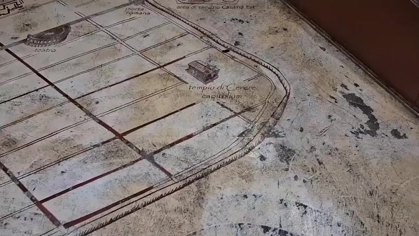 The video shows a close-up, oblique, panning shot of a worn, textured surface displaying a map of an ancient Roman settlement. The map is rendered in a faded brown and red ink with lines and text in Italian. Visible elements include a depiction of a theater, a Roman gate labeled "porta romana," a temple labeled "tempio di Cerere Capitolino," and an area marked "area di servizio Casilina Est" which includes a small circle labeled "voi siete qui" indicating the viewer's current location. There is also an arrow pointing in the direction of Rome, labeled "direzione Roma." The surface itself appears to be stone or a similar material with visible wear and dirt. The background is out of focus, showing a brown, wooden-like edge of the display or enclosure.