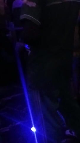 Here is a description of the video:

The video shows a person dancing at what appears to be a party or club. The person is wearing a green shirt and pants and white shoes. They are moving their body and arms to the music. The lighting is dark, with colored spotlights shining on the person and the floor. There are other people in the background, but they are mostly out of focus. The floor is dark and has a textured surface.
