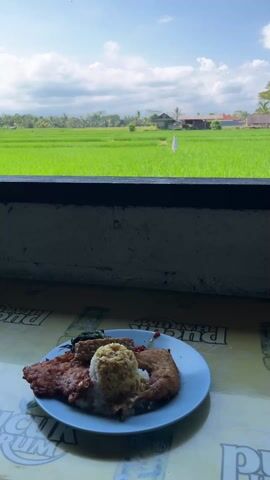 A plate of food rests on a table overlooking a verdant rice paddy. The plate holds a portion of rice, a fried item possibly chicken or cutlet, and other smaller food items that may be vegetables or tofu. The table has a patterned surface with text, mostly obscured, possibly from advertisements or logos. Behind the table, a low wall provides a clear view of the expansive rice field, a couple of houses visible in the distance. The sky is blue with puffy white clouds, suggesting a bright sunny day.