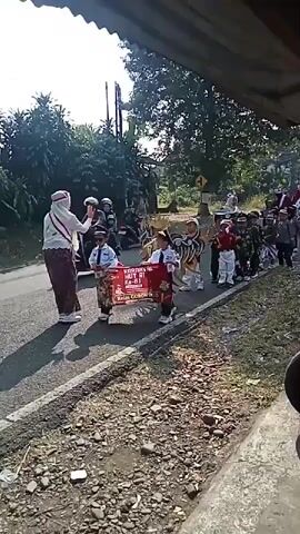 A group of children in costumes and uniforms march down a paved road. The children are of varying ages and appear to be participating in a parade or procession. Some of the children are dressed in traditional Indonesian attire, while others are in military-style uniforms. One child in the front is wearing a large, elaborate costume resembling a dragon or mythical creature. Another child is holding a banner with text in Indonesian. A woman in a headscarf and a sash is walking alongside the children, appearing to be a chaperone or leader. The environment is outdoors, with trees and vegetation visible on the sides of the road. The sun is shining, casting shadows. The road is asphalt and appears to be in a rural or suburban area. There is a motorcycle in the background. The overall scene depicts a lively and festive event involving children.