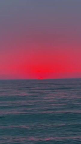 The video shows a sunset over the ocean. The sky is a vibrant red, and the sun is a small, bright orange disc just above the horizon. The ocean is calm, with gentle waves. The colors are saturated and rich, creating a dramatic and beautiful scene.