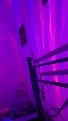 The video shows a static shot of a bedroom. In the foreground is the headboard of a bed, made of dark metal with decorative scrollwork. To the left of the bed is a small doll dressed in a hooded jacket and pants, sitting on a shelf. On the wall behind the doll is a framed picture. The walls are illuminated with a projected light effect that mimics the aurora borealis, cycling through various colors including blue, purple, red, and green. The lighting shifts throughout the video, changing the ambiance of the room.