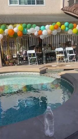 A backyard party is set up around a swimming pool, featuring a balloon arch, inflatable pool floats, chairs, and a DJ setup. The pool is filled with water and has several large inflatable rings floating in it. Some of the rings have animal print patterns. A person wearing shorts and a t-shirt is visible near the edge of the pool. Another person, also dressed casually, is seen standing on the side of the pool. In the background, there are houses, a fence, and some decorations. The sky is clear and blue, indicating a sunny day.
