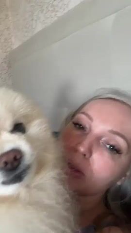 A woman and a small fluffy white dog are lying down together. The dog's face is close to the camera, and its mouth is slightly open, showing its teeth. The woman's face is partially visible behind the dog. They are both in a bed with a patterned wallpaper in the background.